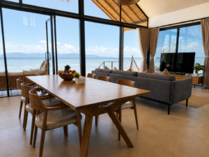 Dining table and grey sofa beside panoramic windows overlooking the sea.