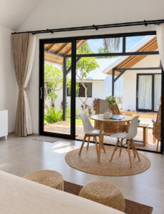 Dining table and chairs beside the glass doors with greenery outside and the bed in the foreground.