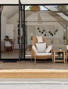 Glass-fronted villa and wooden terrace with a woven chair and side table.
