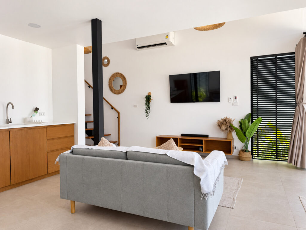 Wide view of the bright living room with a grey sofa, television, kitchen area and staircase.
