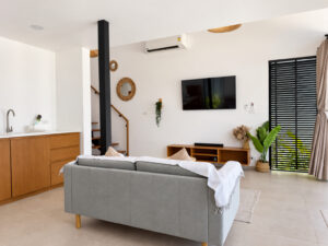 Wide view of the bright living room with a grey sofa, television, kitchen area and staircase.