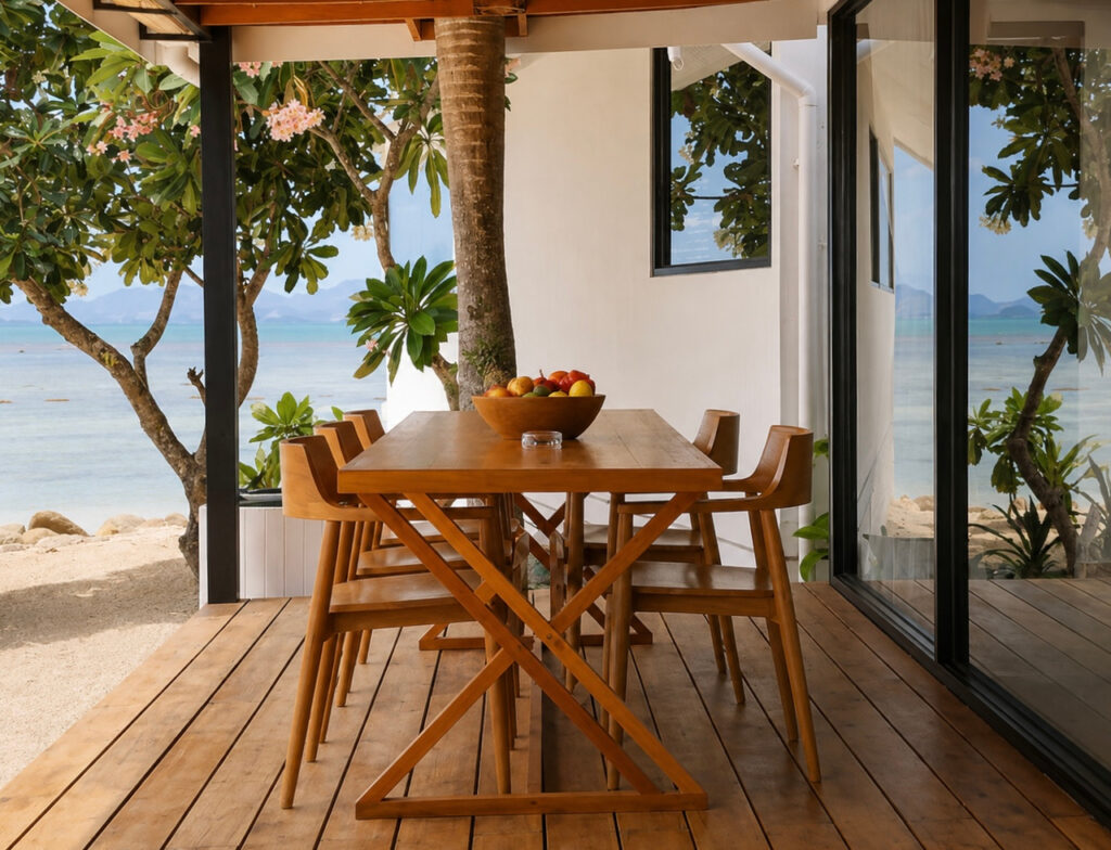 Wooden terrace dining set beside a flowering tree with sand and sea beyond.