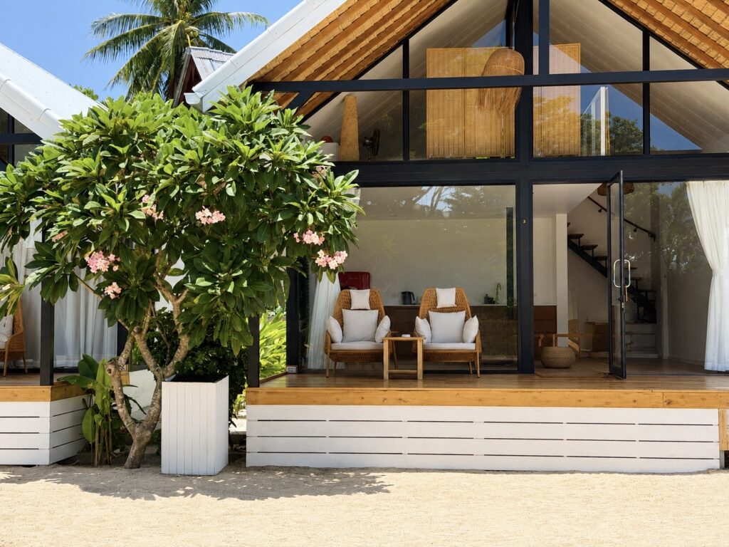 Front view of the glass-fronted villa with terrace chairs, wide steps and a leafy tree.