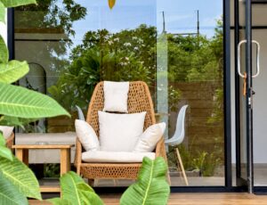 Woven terrace chair with white cushions beside glass doors and leafy plants.