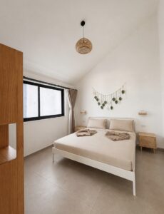 Double bed with folded towels beneath leaf wall decor and a woven pendant light.
