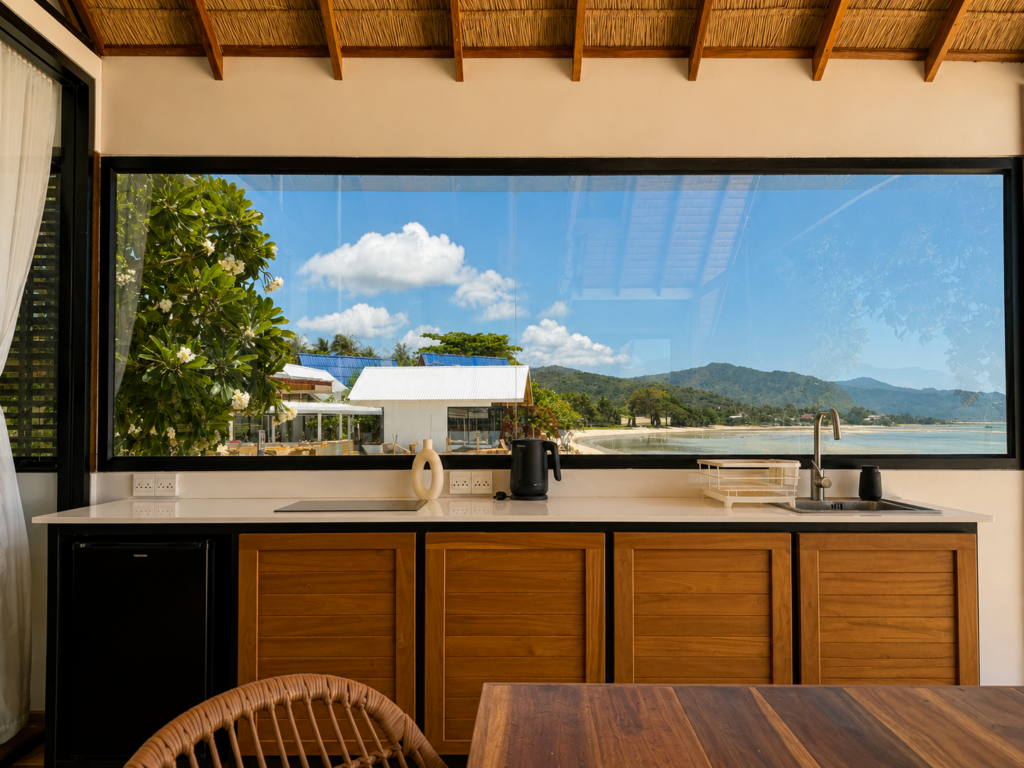 Wooden kitchen cabinets, white counter and sink facing the sea through wide glazing.