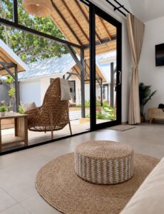 View past a woven pouffe towards the tall glass doors and terrace lounge chairs.