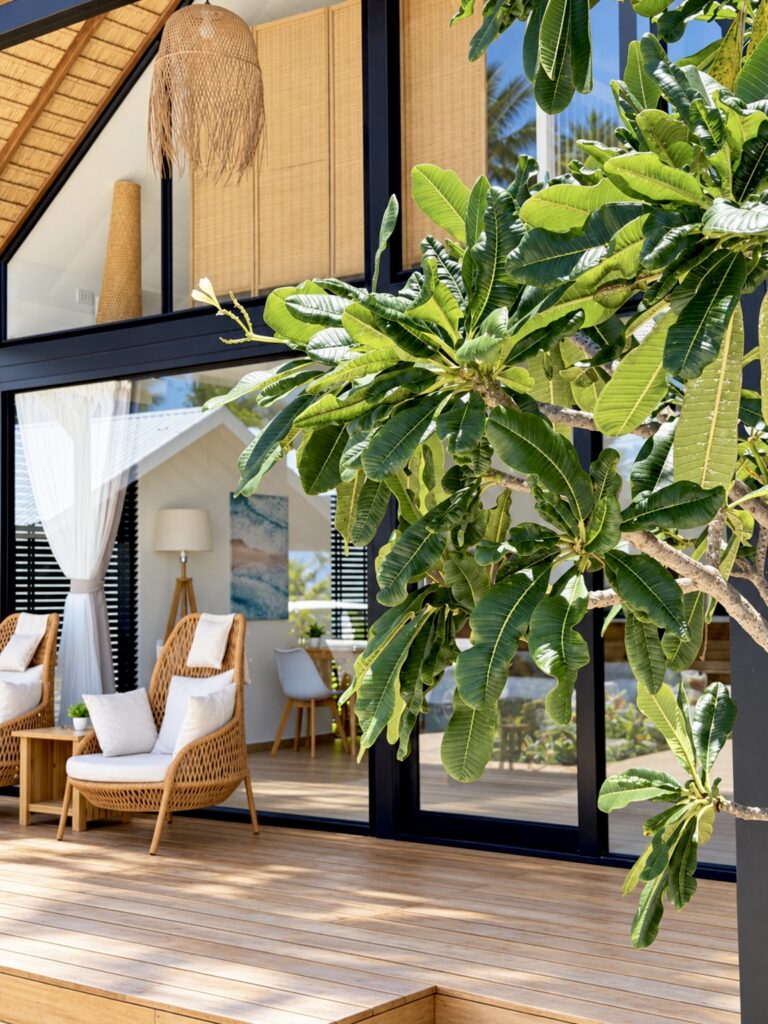 Glass-fronted villa with a wooden terrace, outdoor chairs and surrounding greenery.