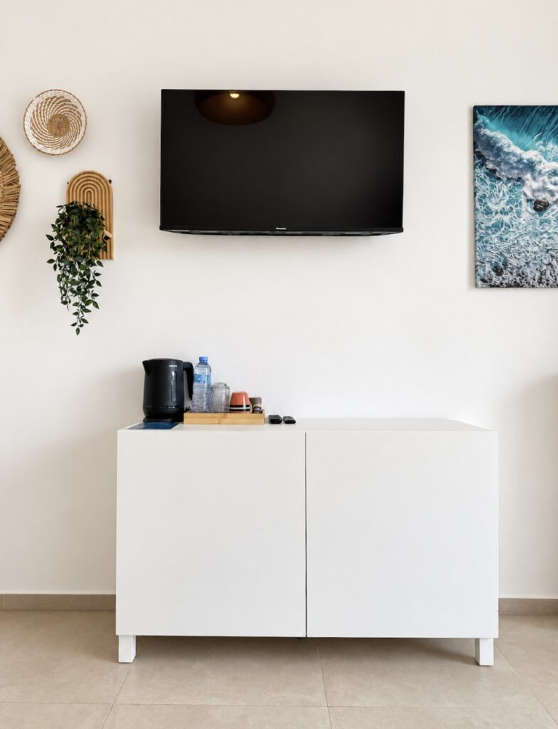 Television and coastal artwork above a white cabinet with an electric kettle and cups.