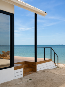 Side view of the villa with wooden terrace steps, sandy beach and sea.