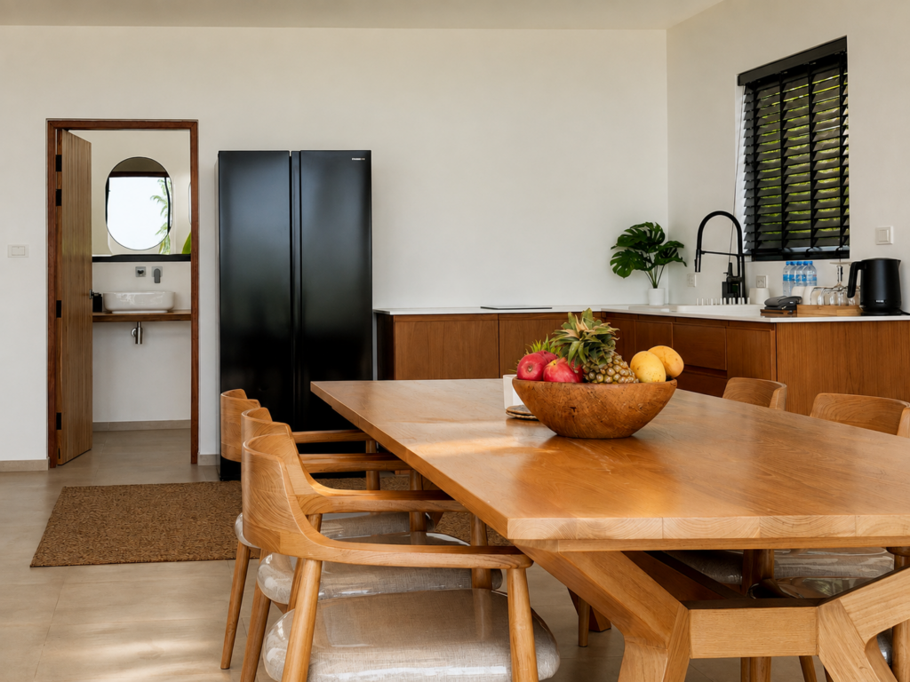 Kitchen area with a refrigerator, sink and wooden dining table with a fruit bowl.