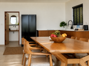 Kitchen area with a refrigerator, sink and wooden dining table with a fruit bowl.