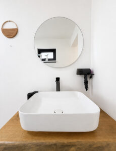 Countertop basin on a wooden shelf beneath a round mirror and hair dryer.