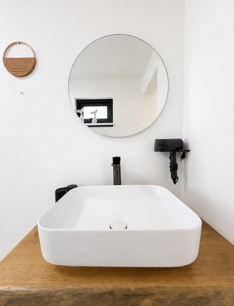 Countertop basin on a wooden shelf beneath a round mirror and hair dryer.