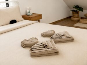 Folded towels on a light-coloured bed beside a wooden bedside table.