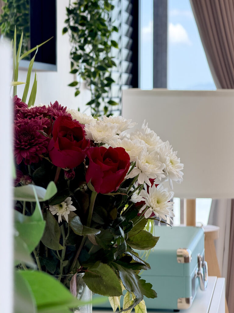 Red and white flowers beside a floor lamp and a mint-coloured case on a sideboard.