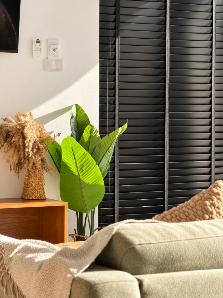 Sofa cushions and indoor plants beside dark window shutters.