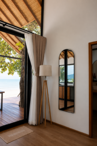Tall wall mirror and tripod floor lamp beside doors opening towards the sea.