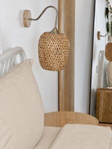 Woven bedside lamp beside light-coloured pillows and a wooden table.