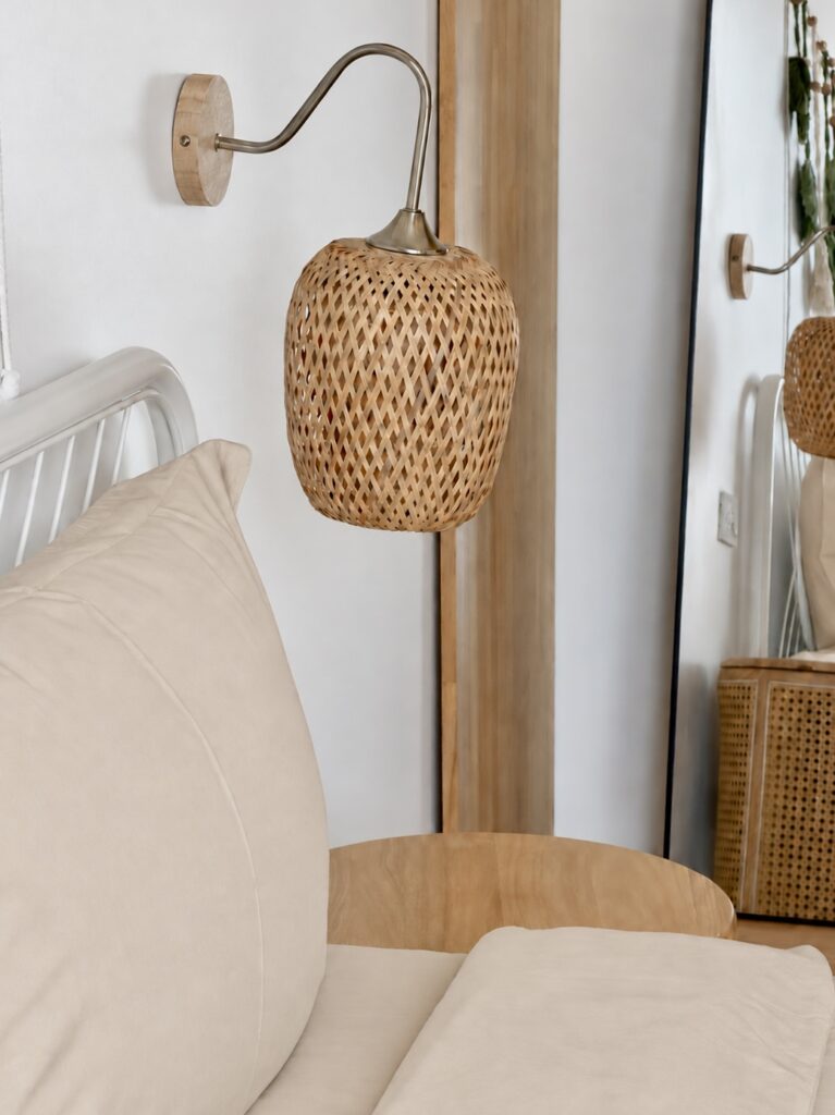 Woven bedside lamp beside light-coloured pillows and a wooden table.